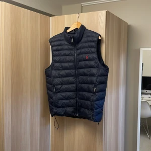 Mörkblå dunväst från Polo Ralph Lauren - Snygg mörkblå dunväst från Polo Ralph Lauren med broderad röd logga på bröstet. Västen har dragkedja framtill och praktiska fickor med dragkedjor. Perfekt för sommaren/våren priset kan sänkas vid snabb affär