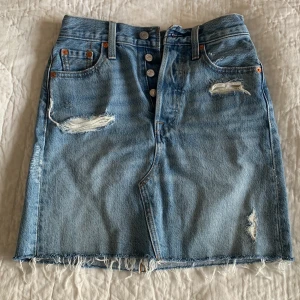 Blå jeanskjol från Levi's - Säljer en snygg blå jeanskjol från Levi's med slitna detaljer och fransig kant. Kjolen har en knappgylf och klassiska femficksdesign. Perfekt till en vit topp för riktiga MammaMia vibes🦋