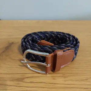 Snyggt flätat bälte i blått, svart och grått med bruna läderdetaljer. Perfekt för att ge din outfit en stilfull touch. Bältet har ett silverfärgat spänne som ger en modern känsla.
