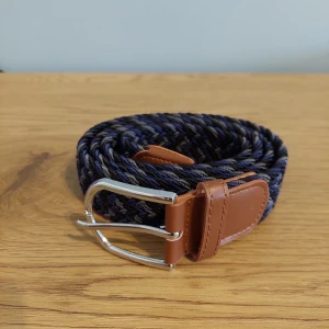 Flätat bälte med läderdetaljer - Snyggt flätat bälte i blått, svart och grått med bruna läderdetaljer. Perfekt för att ge din outfit en stilfull touch. Bältet har ett silverfärgat spänne som ger en modern känsla.