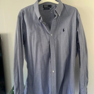 Blå skjorta från Polo - Säljer en stilren blå skjorta från Polo i 100% bomull. Skjortan har en klassisk krage och knappar framtill. Den är långärmad och har en broderad logotyp på bröstet. Perfekt för en snygg och avslappnad look.