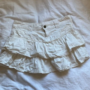 Vit volangkjol från Bershka - Säljer en söt vit kortkjol från Bershka med volanger. Perfekt för varma dagar och ger en lekfull touch till din outfit. Kjolen har en knapp och dragkedja framtill.