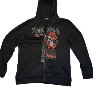 Svart hoodie med dödskallemotiv från Rock Rebel - Säljer en svart hoodie från Rock Rebel med ett coolt dödskallemotiv och textdetaljer i rött och grått. Hoodien har en dragkedja med en unik dödskalle-detalj och en stor ficka framtill. Perfekt för dig som gillar en rockig stil! 🖤