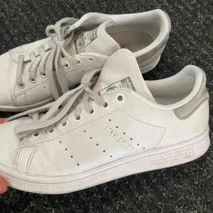 Vita Adidas Stan Smith sneakers - Säljer ett par klassiska vita Adidas Stan Smith sneakers. Skorna har en stilren design med snörning och perforerade detaljer på sidorna. Perfekta för en avslappnad och trendig look.