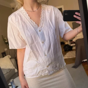 Vit blus med spetsdetaljer - Säljer en söt vit blus med korta ärmar och v-ringning. Blusen har fina spetsdetaljer längs halsringningen och knappar framtill. Perfekt för en somrig look!