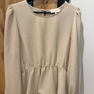 Beige blus från ONLY - Säljer en stilren beige blus från ONLY. Blusen har långa ärmar och en lätt rynkad midja som ger en fin siluett. Perfekt för en elegant look. Den har en knappdetalj i nacken för enkel påklädning.