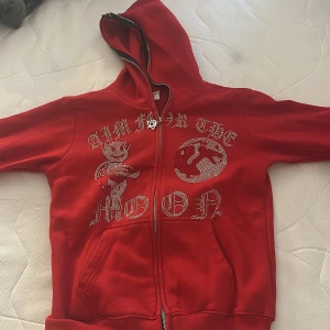 Röd huvtröja med strassdetaljer i storlek s  - Snygg röd huvtröja med dragkedja och strassdetaljer som bildar texten 'Aim for the Moon'. Tröjan har en huva och ribbade muddar vid ärmslut och nederkant. Perfekt för en cool och avslappnad stil.