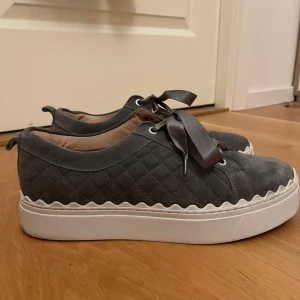 Grå sneakers - Snygga grå sneakers med quiltad design och en dekorativ satinrosett. Skorna har en rund tå och en vit sula med vågig kant. Perfekta för en stilren look.