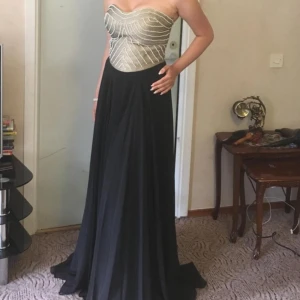 festklänning balklänning  - Elegant axelbandslös klänning med en glittrig guldfärgad överdel och en lång, flödande svart kjol. Perfekt för en festlig kväll. Använd under en kväll. Köpt för 3000kr