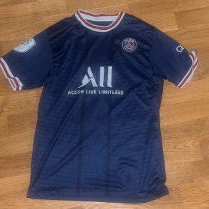 Blå PSG fotbollströja - Säljer en blå PSG fotbollströja med Neymar Jr och nummer 10 på ryggen. Tröjan har korta ärmar och vita detaljer. Tillverkad i 100% mikrofiber för en lätt och bekväm känsla. Perfekt för fotbollsfans!