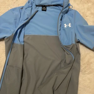 Blå och grå vindjacka från Under Armour  - Säljer en stilren blå och grå vindjacka från Under Armour. Jackan har en dragkedja framtill och en praktisk huva. Perfekt för utomhusaktiviteter och träning.