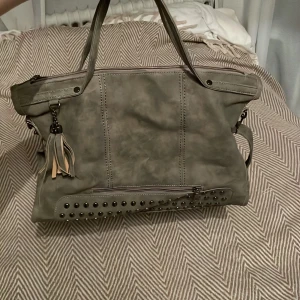 Grå axelväska med nitar - Snygg grå axelväska med dekorativa nitar och tofsdetalj. Väskan har både handtag och en justerbar axelrem för flexibilitet. Den är rymlig med dragkedja upptill och en extra ficka med dragkedja på baksidan. Perfekt för att bära med sig allt man behöver.