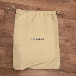 Skopåse från Axel Arigato - Praktisk skopåse från Axel Arigato i beige med dragsko. Perfekt för att förvara och skydda dina skor. Enkel och stilren design med märkets logotyp tryckt i svart. Kan även användas som en idrotts påse eller ha plånbok o smycken i när man reser!!!!