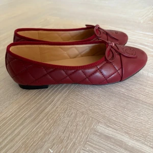 Vinröda ballerinaskor ch*nel - Snygga vinröda ballerinaskor med quiltad design och dekorativ rosett på tån. Skorna är i skinn. Storlek 38 tyvärr förstora för mig. Kan skicka original bilder om intresserad