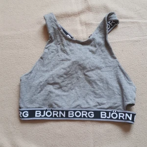 Grå linnetopp från Björn Borg - Säljer en grå linnetopp från Björn Borg med svarta och vita elastiska band med logotyp. Toppen har korsade band i ryggen och är perfekt för en sportig look. Lite slitage på banden (se bilder), därav priset
