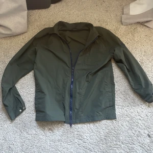 Grön windbreaker från Knowledge Cotton Apparel - Snygg grön vindjacka från Knowledge Cotton Apparel med dragkedja framtill. Jackan har en klassisk krage och långa ärmar med knappdetaljer vid ärmsluten. Perfekt för blåsiga dagar.