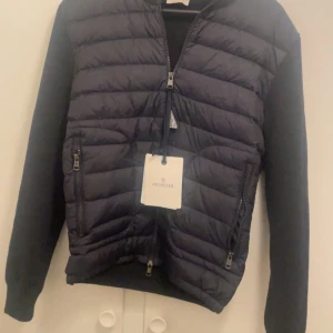 Navy blue Moncler jacka  - Säljer en Snygg Navy blue quiltad jacka från Moncler som är helt oanvändbar utan luva som är  med dragkedja framtill och långa ärmar. Jackan har en stilren design med Moncler-logga på ärmen och praktiska fickor med dragkedja. Perfekt för vårdagarna samt sommardagar.