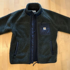 Grön fleecejacka från Carhartt - Säljer en grön fleecejacka från Carhartt med dragkedja och bröstficka. Jackan har en mysig och varm design, perfekt för kyligare dagar. Den har långa ärmar och en praktisk dragkedja framtill.