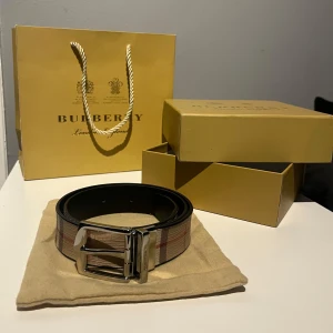  Burberry bälte - Snyggt Burberry bälte med klassiskt rutigt mönster i beige och bruna toner. Bältet har en stilren silverfärgad spänne och kommer med påse och låda. Perfekt för att ge en touch av lyx till din outfit.