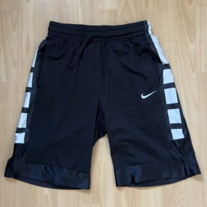 Svarta sportshorts från Nike - Snygga svarta sportshorts från Nike med vita detaljer längs sidorna. De har en bekväm passform med snörning i midjan och är perfekta för träning. Den ikoniska Nike-loggan syns på ena benet.