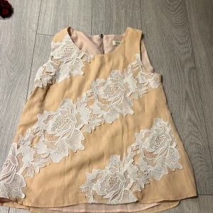 Beige ärmlös blus med spetsdetaljer - Säljer en elegant beige ärmlös blus från JOVONISTA med vackra vita spetsdetaljer i blommönster. Blusen har en dragkedja i ryggen för enkel på- och avtagning. Perfekt för en stilren look.