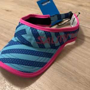 Keps/ run cap från Salming - Snygg solskärm från Salming i blått med rosa detaljer. Perfekt för soliga dagar och sportaktiviteter. Justerbar passform för optimal komfort. Helt ny!