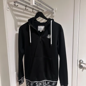 Svart hoodie från SikSilk - Säljer en stilren svart hoodie från SikSilk med vit logga på bröstet och vita snören i huvan. Tröjan har en dragkedja och ribbad kant med SikSilk-mönster vid ärmslut och nederkant. Perfekt för en avslappnad stil.