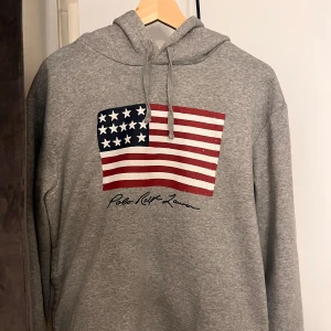 Polo ralph lauren hoodie grå Strlk M amerikansk flagga - Säljer en grå hoodie från Polo Ralph Lauren med en stor amerikansk flagga på framsidan. Köpt i affär och den är knappt använd!
