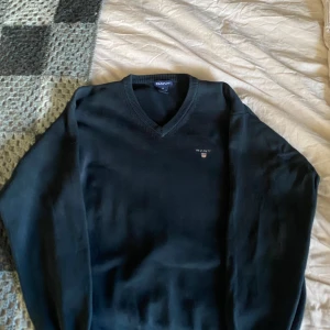 Tröja  från GANT - Säljer en stilren mörkblå tröja från GANT med v-ringning och broderad logotyp på bröstet. I storlek M.Tröjan har långa ärmar och är perfekt för en klassisk look.