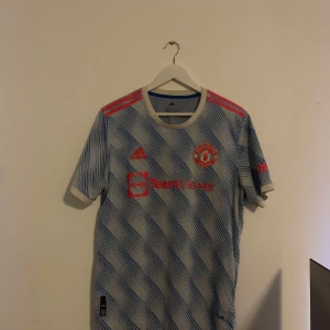 Blå och vit Manchester United fotbollströja från Adidas authentic - Säljer en blå och vit randig fotbollströja från Adidas med korta ärmar. Tröjan har röda detaljer och ett tryck på framsidan.  Perfekt för fotbollsfans! Nr18 Bruno storlek L