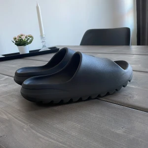 Yeezy slides  - Säljer ett par svarta yeezy slides från Adidas med en stilren design. De har en väldigt mjuk känsla och är enligt mig en av de skönaste tofflorna på marknaden. Jag har använt tofflorna ungefär 10 gånger så de är fortfarande i prima condition. Säljs för ett sjukt bra pris (fraktar samma dag som köp). Hör av er vid funderingar!