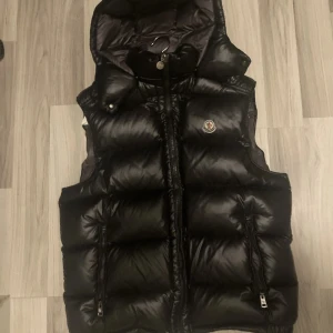 Svart dunväst med huva - Snygg svart dunväst från Moncler med huva och dragkedja. Västen har en glansig yta och två fickor med dragkedjor framtill. Perfekt för kyligare dagar. Storlek L/M
