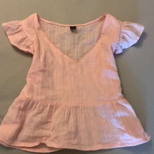 Rosa blus från Shein - Söt rosa blus från Shein med v-ringning och korta ärmar. Blusen har en lätt volangdetalj vid nederkanten som ger en feminin touch. Perfekt för varma dagar!