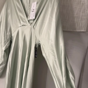 Ljusgrön långklänning i satin - Elegant ljusgrön långklänning i satin med v-ringad design. Klänningen har långa ärmar och en smickrande passform som ger en stilren look. Perfekt för speciella tillfällen.
