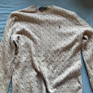 Beige stickad tröja från Ralph Lauren - Säljer en klassisk beige stickad tröja från Ralph Lauren med kabelstickat mönster och broderad logotyp på bröstet. Tröjan har långa ärmar och en rund halsringning. Perfekt för en stilren look!