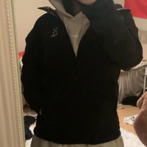 Svart vindjacka från Haglöfs - Säljer en svart vindjacka från Haglöfs med dragkedja och huva. Perfekt för blåsiga dagar och har en stilren design. Jackan har långa ärmar och är tillverkad i ett slitstarkt material. Använd 4 gånger och vattentätt.