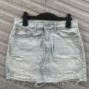 Mini Ljusblå jeanskjol med slitningar - Trendig ljusblå jeanskjol med slitna detaljer och rå kant. Kjolen har klassiska fem fickor och knappar