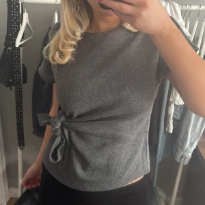 Grå ribbad topp med knyt - Säljer en stilren grå ribbad topp med korta ärmar och knytning i midjan. Perfekt för en avslappnad men chic look. 
