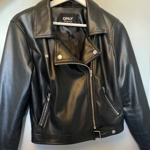Svart skinnjacka från ONLY - Snygg svart skinnjacka från ONLY med dragkedja och knappar. Jackan har en klassisk bikerstil med bälte i nederkant och två fickor med dragkedja. Perfekt för en cool look.
