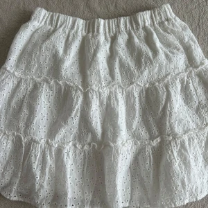 Vit kjol med spetsdetaljer - Charmig vit kjol med spetsdetaljer och volanger. Perfekt för en somrig look. Den har en elastisk midja för bekväm passform.