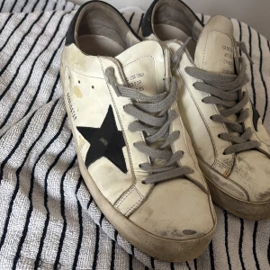 Vita sneakers med svart stjärna - Snygga vita sneakers från Golden Goose med en ikonisk svart stjärna på sidan. Skorna har grå snörning och en vintage-look. Perfekta för en avslappnad stil. Lite heeldrag på hälarna, men lagat med lim vet ej hur länge limet håller men kan lagas hos skomakare sick:5/10 Storlek 43 passar även 44