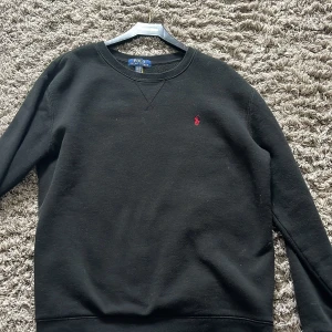 Svart tröja från Ralph Lauren - Säljer en klassisk svart tröja från Ralph Lauren med rund halsringning och ett litet rött broderat märke på bröstet. Perfekt för en stilren look. 