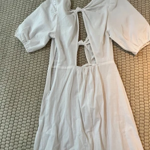 Vit klänning med öppen rygg och knyt - Säljer en stilren vit klänning med korta puffärmar och en elegant öppen rygg. Klänningen har knytning framtill och baktill vilket ger en unik look. Perfekt för sommarens alla tillfällen!
