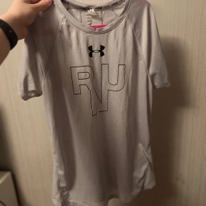 Grå tränings-t-shirt från Under Armour - Säljer en grå tränings-t-shirt från Under Armour med texten 'RUN' på framsidan. T-shirten är kortärmad och tillverkad i ett lätt och andningsbart material, perfekt för löpning och träning.