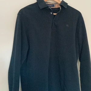 Svart långärmad pikétröja från Burberry - Säljer en stilren svart långärmad pikétröja från Burberry. Knapparna är lite utslitna men annars är den i bra form. Hör av er om ni har fler frågor!