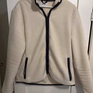 Vit fleecejacka från Easy Wear - Säljer en mysig vit fleecejacka från Easy Wear med marinblå detaljer. Jackan har en dragkedja framtill och två praktiska fickor med dragkedjor. Perfekt för kyliga dagar!