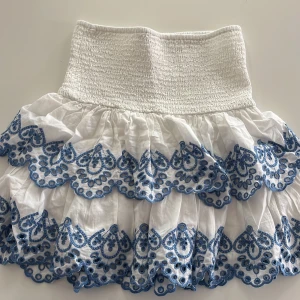 Vit kjol med blå spetsdetaljer - Charmig vit kjol med vackra blå spetsdetaljer. Den har en elastisk midja som ger en bekväm passform. Perfekt för en somrig look! Aldrig använd 