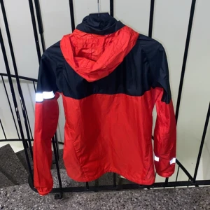 Röd och svart vindjacka från Nike - Säljer en snygg röd och svart vindjacka från Nike med huva och dragkedja. Jackan har reflekterande detaljer på ärmarna och är perfekt för utomhusaktiviteter. Den är lätt och bekväm att bära.köpte den för 1000kr de står även på lappen insidan. Helt ny 