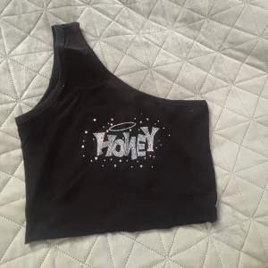 Svart oneshoulder topp med text - Snygg svart oneshoulder topp med texten 'HONEY' i glittriga stenar på framsidan. Perfekt för en trendig look.