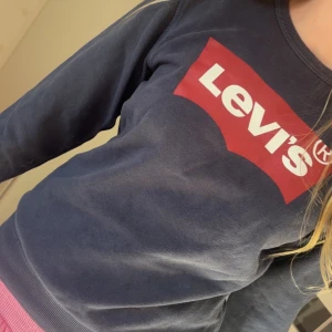 Mörkblå sweatshirt från Levi's - Säljer en mörkblå sweatshirt från Levi's med det klassiska röda och vita logotyptrycket på framsidan. Tröjan har långa ärmar och ribbade muddar vid ärmslut och nederkant. Perfekt för en avslappnad stil.